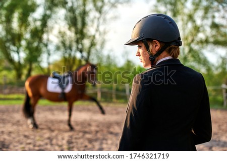 
A horse rider girl stands on a farm wearing a helmet on his head. In the background stands a horse raised his leg.
