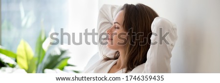 Beautiful 30s woman closed eyes put hands behind head relaxing on comfortable sofa in cozy warm light living room with houseplants, no stress concept, horizontal photo banner for website header design Royalty-Free Stock Photo #1746359435