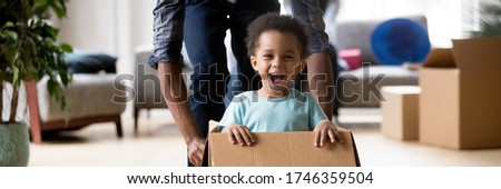 African dad play with excited toddler son riding seated inside of box in moving day at new home, little kid enjoy game with daddy, relocation concept. Horizontal photo banner for website header design