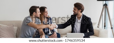 Happy couple young family flat renters shake hands with realtor seated on sofa in living room. Make real estate deal, first dwelling purchase concept. Horizontal photo banner for website header design