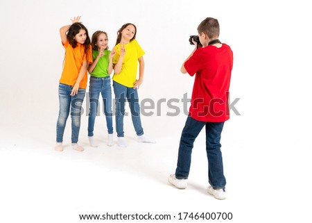 A boy photographer takes pictures of three cute girls in bright T-shirts on a white background in the studio. Young photographer and blogger.