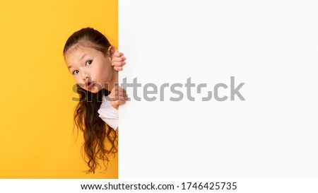 Funny asian girl hiding behind blank white advertising billboard with copy space and peeking out, yellow studio wall
