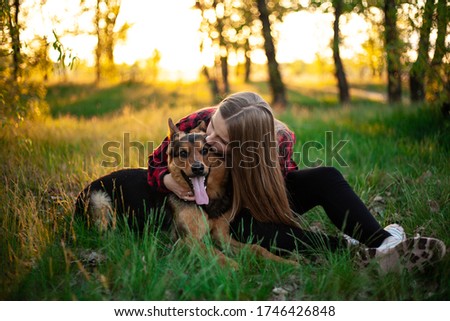 Happy blonde girl is played with a dog. Girl hugs a dog sitting on the grass. Dog Shepherd, female. Friend dog. Outdoor photo shoot at sunset
