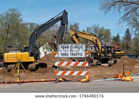 Road Closed sign with heavy construction equipment in the background framed by trees and a blue sky.