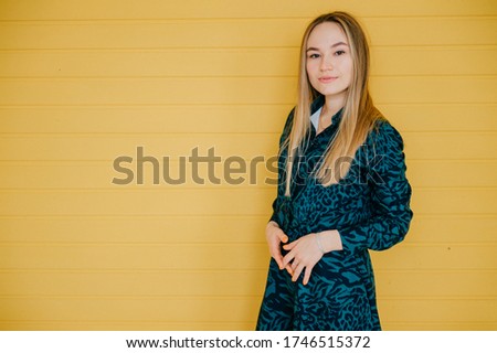 Cheerful caucasian female with long fair hair in green dress smiles, picture isolated on yellow background