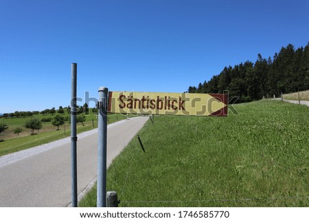 Beige and Brown sign in German pointing the way to a scenic view of the a mountain in Switzerland. A road, grass and trees are in the background.