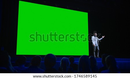 Software Startup Founder Does Presentation of New Product to the Audience, Behind Him Movie Theater with Green Screen, Mock-up, Chroma Key. Business Conference Live Event or Device Reveal