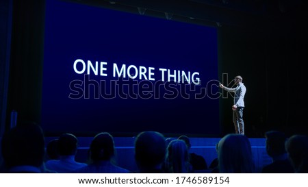 Live Event with Brand New Products Reveal: Keynote Speaker Presents New Device to Audience. Movie Theater Screen Shows Text -one more thing- Royalty-Free Stock Photo #1746589154