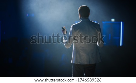 On Stage: Successful Motivational Speaker, Talking about Happiness, Self, Success, Empowerment, Efficiency, Health, and How to Be More Productive. Large Conference Hall with Cinematographic Light Royalty-Free Stock Photo #1746589526