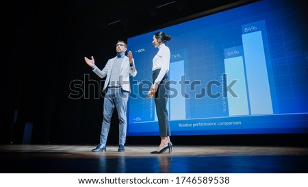 On Stage, Successful Female CEO and Male COO Speakers Present Company's New Product, Show Infographics, Statistics on Big Screen, Talk About Growth. Live Event, Tech Startup, Business Conference Royalty-Free Stock Photo #1746589538