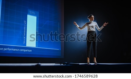 On-Stage Successful Female Speaker Presents Technological Product, Uses Remote Control for Presentation, Showing Infographics, Statistics Animation on Screen. Live Event / Device Release. Royalty-Free Stock Photo #1746589628