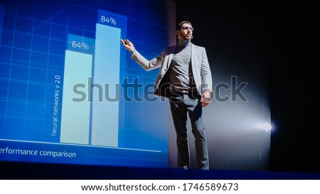 On-Stage Speaker Gives Presentation of Technological Device, Shows Infographics Animation on Big Screen. Auditorium Hall Live Event, Product Release, Start-up Conference. Low Angle Royalty-Free Stock Photo #1746589673