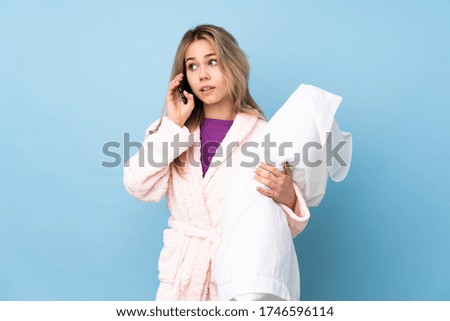 Teenager Russian girl in pajamas isolated on blue background keeping a conversation with the mobile phone with someone