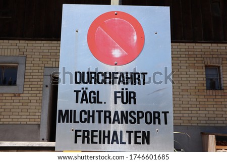 White sign with a red circle which says in German: Keep driveway free every day for milk transport. A brick wall with Windows is in the background.