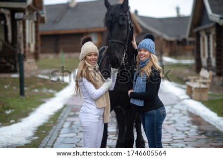 Picture of two young attractive sisters in warm jackets, trousers, boots, scarfs and knitted hats with a horse outside in winter