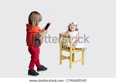 A boy of 3 years old photographs a baby,  Using a smartphone. Girl poses funny to him on a chair. Studio light. Isolated. Lifestyle. Friendship concept, siblings, mobile photo. Horizontal. Caucasian.