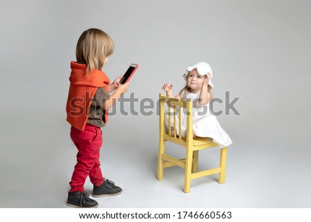 A boy of 3 years old photographs a baby,  Using a smartphone. Girl poses funny to him on a chair. Studio light. Isolated. Lifestyle. Friendship concept, siblings, mobile photo. Horizontal. Caucasian.