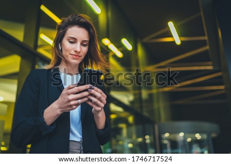 Successful female banker using smartphone outdoors while standing near office background yellow neon lights, young woman professional manager working on mobile device near skyscraper at night