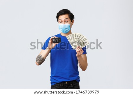 Money, covid-19, easy payment, investment and banking concept. Excited and surprised asian guy staring at credit card, place cash on deposit, holding dollars, standing in medical mask