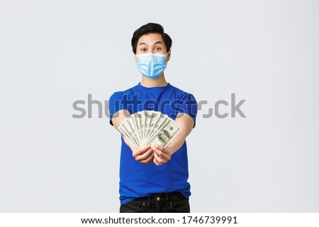 Money, lifestyle, insurance and investment concept. Look how much cash I got. Asian excited guy in medical mask show-off or bragging, showing dollars to you, standing grey background