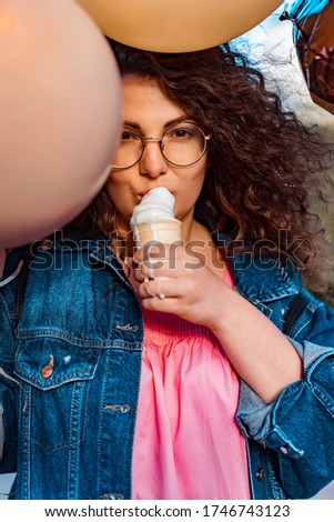 Young beautiful brown curly hair woman in pink long dress with colorful yellow, golden and black balloons and vanilla ice cream cone is walking down the street going to the celebrate a Birthday party