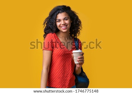 Half-length portrait of cute smiling mixed race tourist with a backpack holding paper cup of coffee. Isolated over yellow background. 