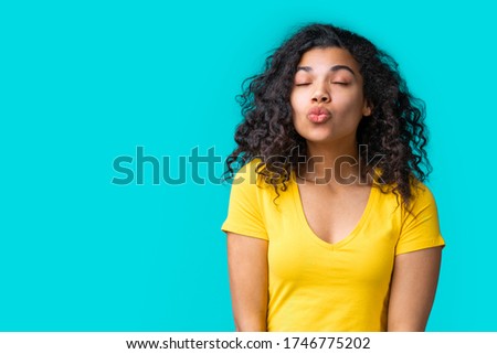 Studio shot of beautiful dark skinned girl in simple basic bright colored yellow t-shirt giving a kiss isolated over blue background. Copy space for your text.