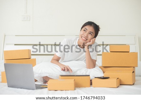 An Asian woman is using a tablet and a computer to happily find and choose products in her bed at home. Around her there are a lot of boxes ordered from the online website. This photo have copy space