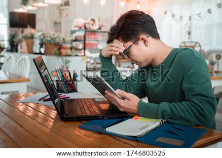 Asian male students feel tired from working with computers. A young businessman uses a laptop to sit at the wooden table of a modern coffee shop. Freelance Translator Working With A Laptop Royalty-Free Stock Photo #1746803525