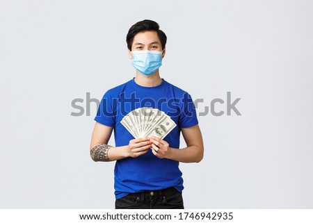 Money, lifestyle, insurance and investment concept. Handsome satisfied asian man earned lots cash, showing dollars and smiling pleased in medical mask, got rich