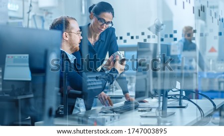 Modern Factory: Female Project Supervisor Talks to a Male Industrial Engineer who Works on Computer. They Inspect Machinery Mechanism and Design Improved Version. Working High-Tech Industrial Facility Royalty-Free Stock Photo #1747003895