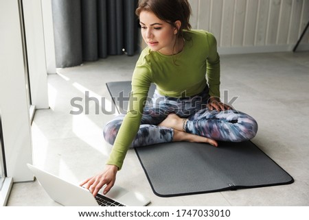 Photo of focused brunette sportswoman using laptop while working out on mat in cozy room