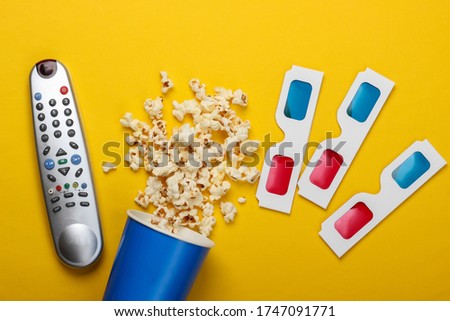 Movie time. Cardboard bucket of popcorn, tv remote and Stereoscopic anaglyph disposable paper 3d glasses on yellow background. Top view