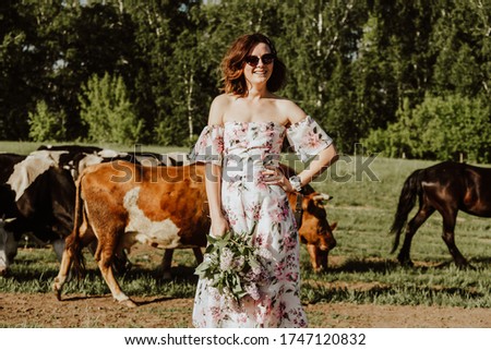 Portrait  outdoor atmospheric lifestyle photo of young beautiful  darkhaired woman in a  pink dress  stands on the background of a herd of cows. Girl calling with herself on a journey
