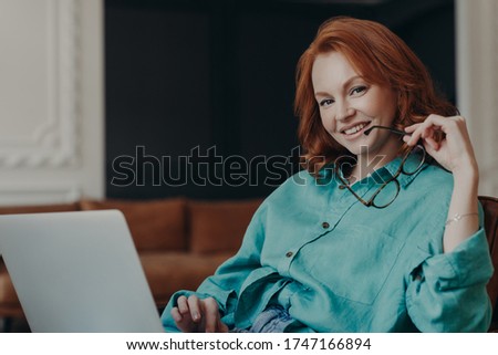 Positive good looking young woman freelancer works remotely on laptop computer, connected to wireless free internet at home, holds glasses, dressed in shirt, smiles happily, being in good mood