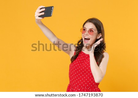 Excited young brunette woman girl in red summer dress, eyeglasses posing isolated on yellow background. People lifestyle concept. Mock up copy space. Doing selfie shot on mobile phone spreading hands