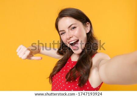 Close up of cheerful brunette woman girl in red summer dress isolated on yellow background. People lifestyle concept. Mock up copy space. Doing selfie shot on mobile phone pointing thumb on herself