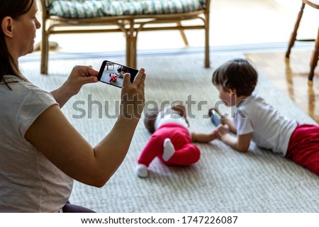 Shot of mother taking photo of baby and son with smartphone at home.Mom making digital family memories with kids.Big brother and baby sister looking at smartphone while mother taking pictures of them.