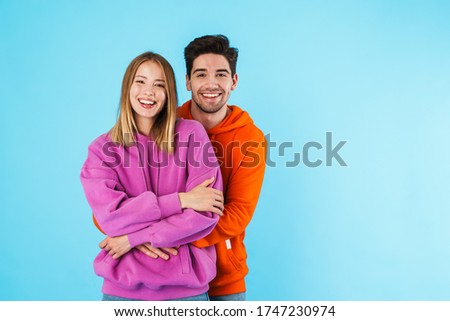 Portrait of a cheerful young couple wearing hoodies standing isolated over blue background, hugging