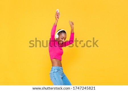 Happy African American woman wearing headphones listening to music and dancing on colorful yellow isolated background