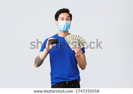 Money, covid-19, easy payment, investment and banking concept. Handsome asian young man assure you need this, showing credit card and cash dollars, wink camera hinting, wear medical mask