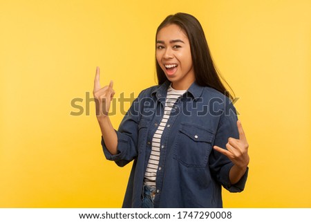 Yeah, that's wonderful! Portrait of joyful girl in denim shirt feeling crazy, showing devil horns, rock and roll hand gesture, delighted of success. indoor studio shot isolated on yellow background