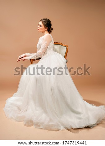 Beautiful bride in white wedding dress sitting in a vintage chair. Studio portrait with brown background. Fashionable wedding gown. 