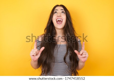 Charming carefree woman with positive expression, points up with both index fingers, dressed in casual clothing, has broad interested smile, isolated over yellow background. Look there, please.