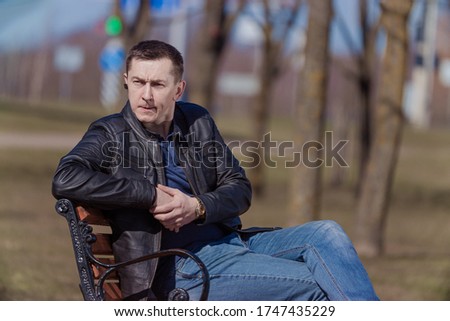 A young adult man in a black leather jacket and jeans sits on a bench in a park on a city street on a sunny day