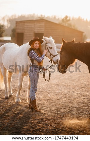 A young cowgirl woman stroking a horse in the sunset at her ranch. Flare effect.