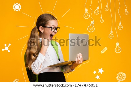 Modern education of children. Overjoyed school girl with backpack is happy with test result, holds laptop in hands, over yellow background in studio