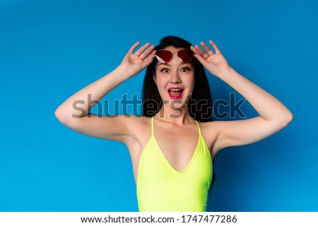 Summer Offer. Excited Asian Girl In Swimsuit Taking Off Sunglasses In Amazement, Yelling Wow, Looking At Camera While Posing Over Blue Background