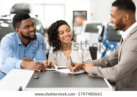 Car Buyers. African Couple Signing Papers With Salesman Standing In Dealership Store. Selective Focus