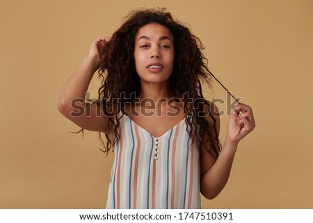 Attractive young long haired curly dark skinned female playing with her hair while looking pensively at camera with light smile, isolated over beige background